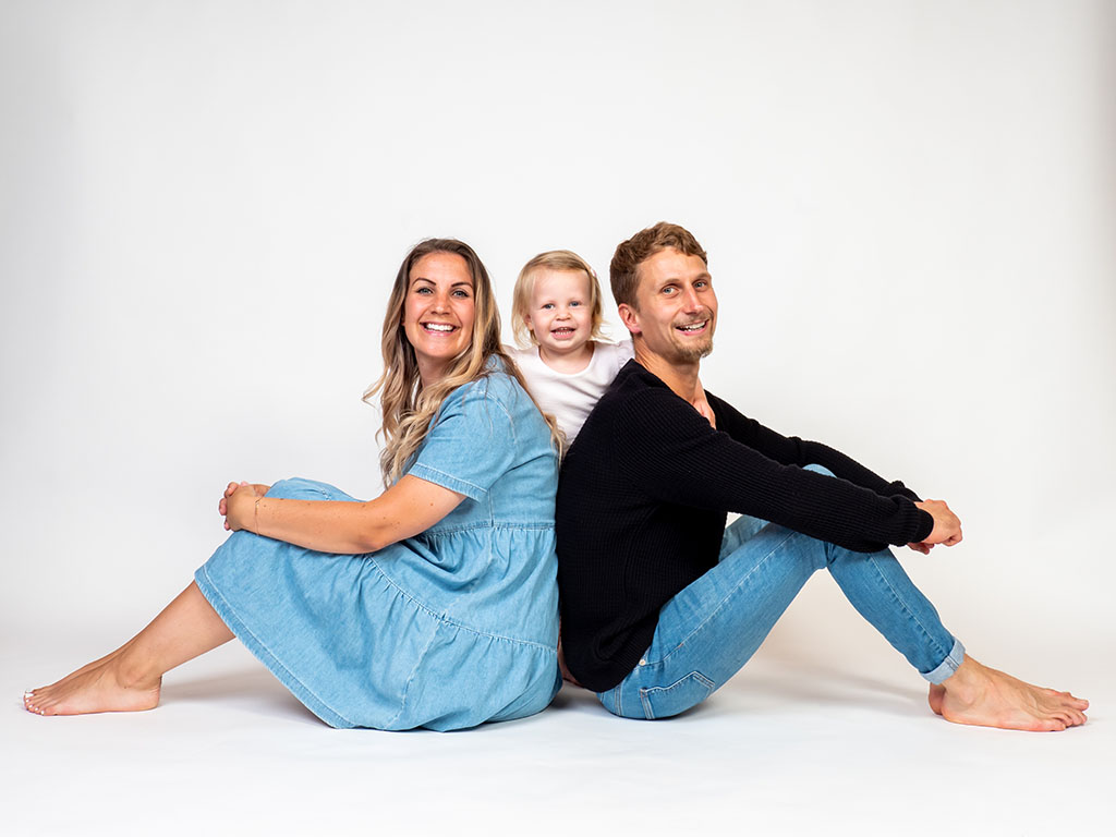 mum and dad sitting on floor back to back with daughter standing between them taken by family photographer in Braintree